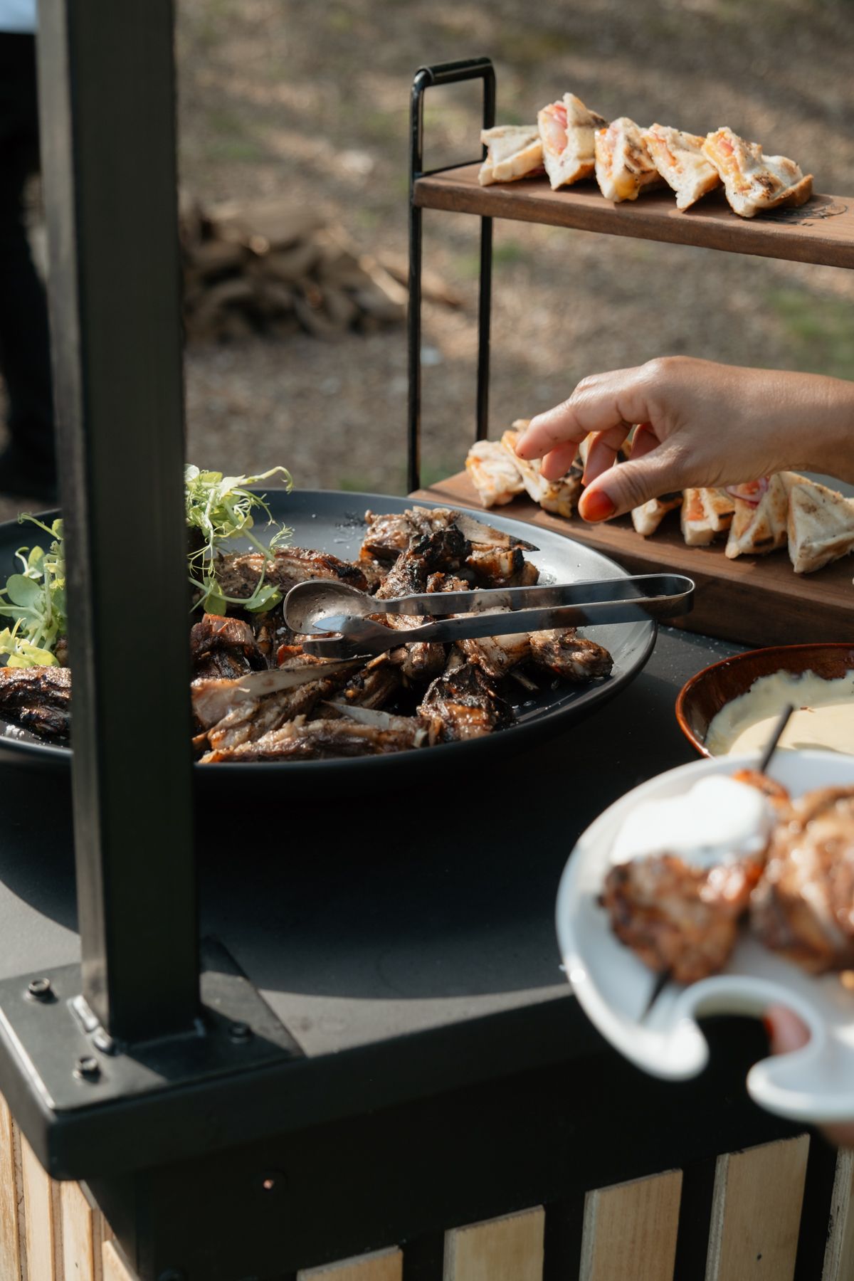 A hand reaches for appetizers on a tiered wooden stand beside a plate of grilled meats at a catered event.