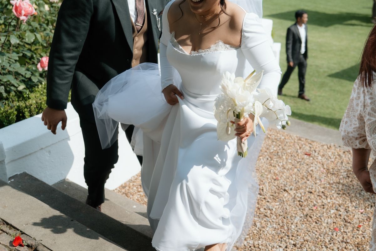 Bride in white long-sleeve gown carrying bouquet walks with groom on garden pathway.