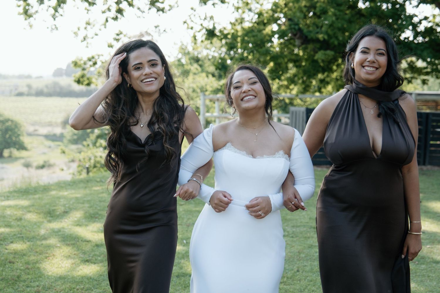 Smiling bride flanked by two bridesmaids in chocolate brown satin dresses on a sunny garden lawn.