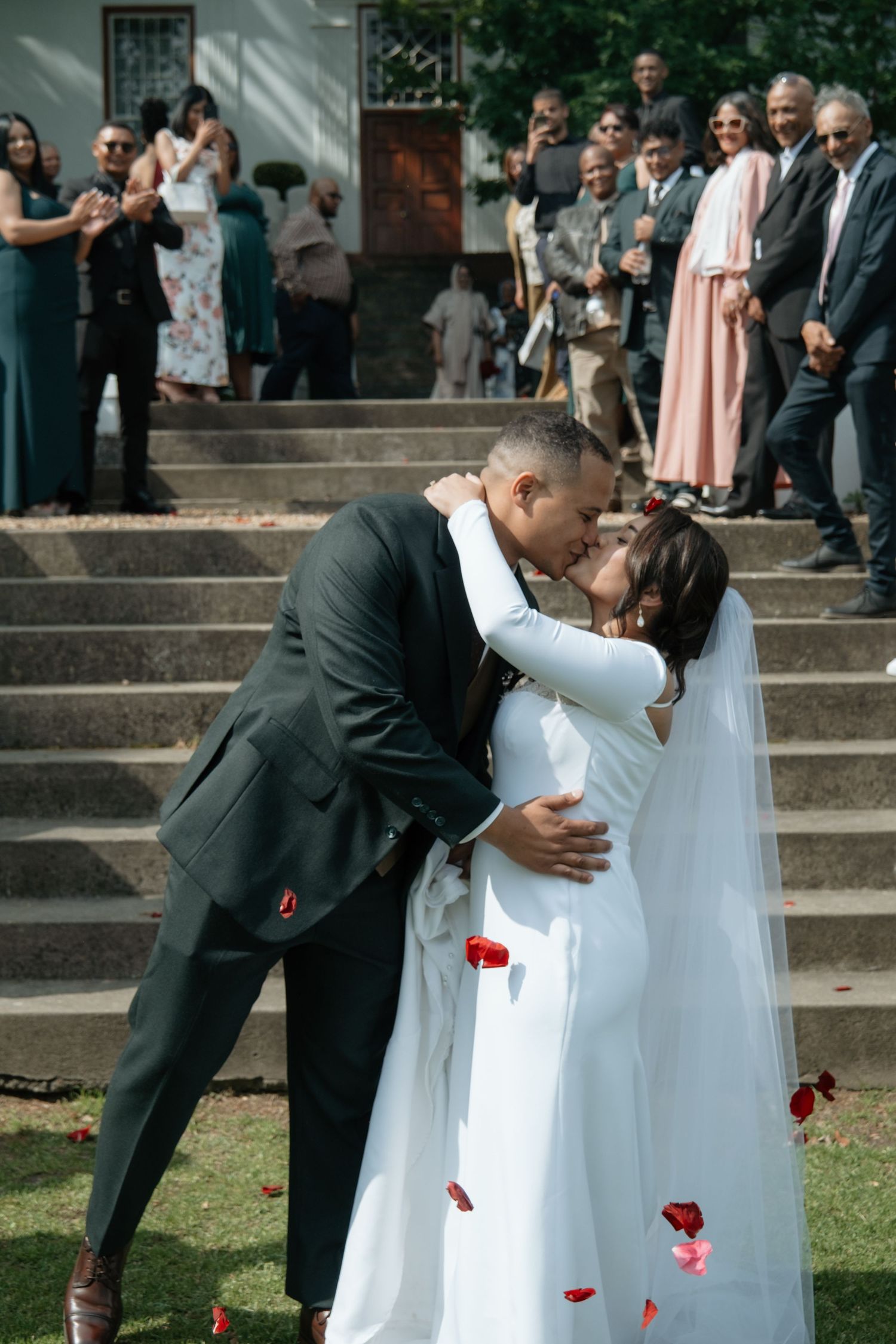 Newlyweds share first kiss surrounded by cheering guests on steps as red rose petals fall around them.