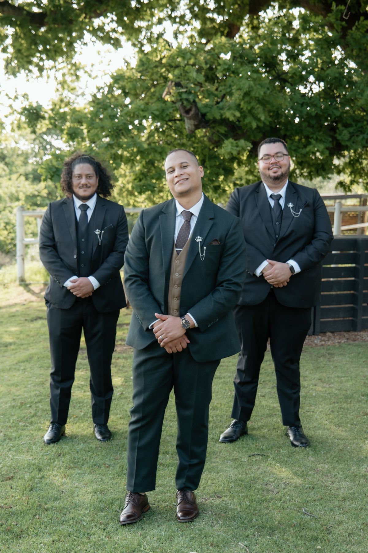 Groom and groomsmen in matching dark suits posing outdoors under large tree at wedding.