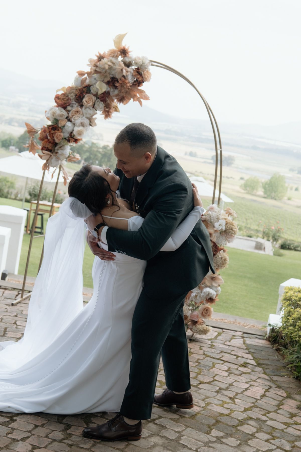 Bride and groom embrace and kiss under circular floral arch at outdoor ceremony.