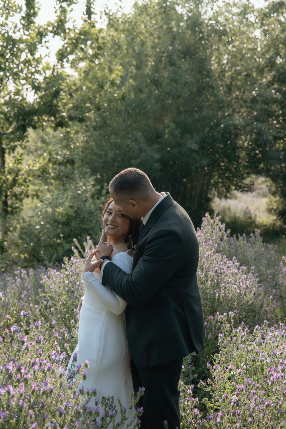 Groom embracing smiling bride from behind in a lush purple lavender garden setting.