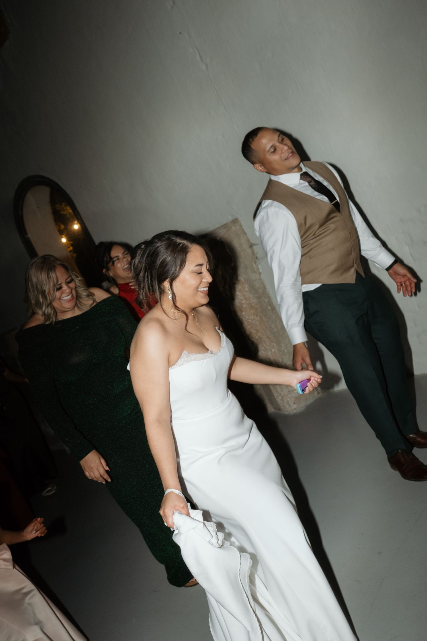 Bride in white strapless gown dances joyfully with wedding party guests at lively reception.