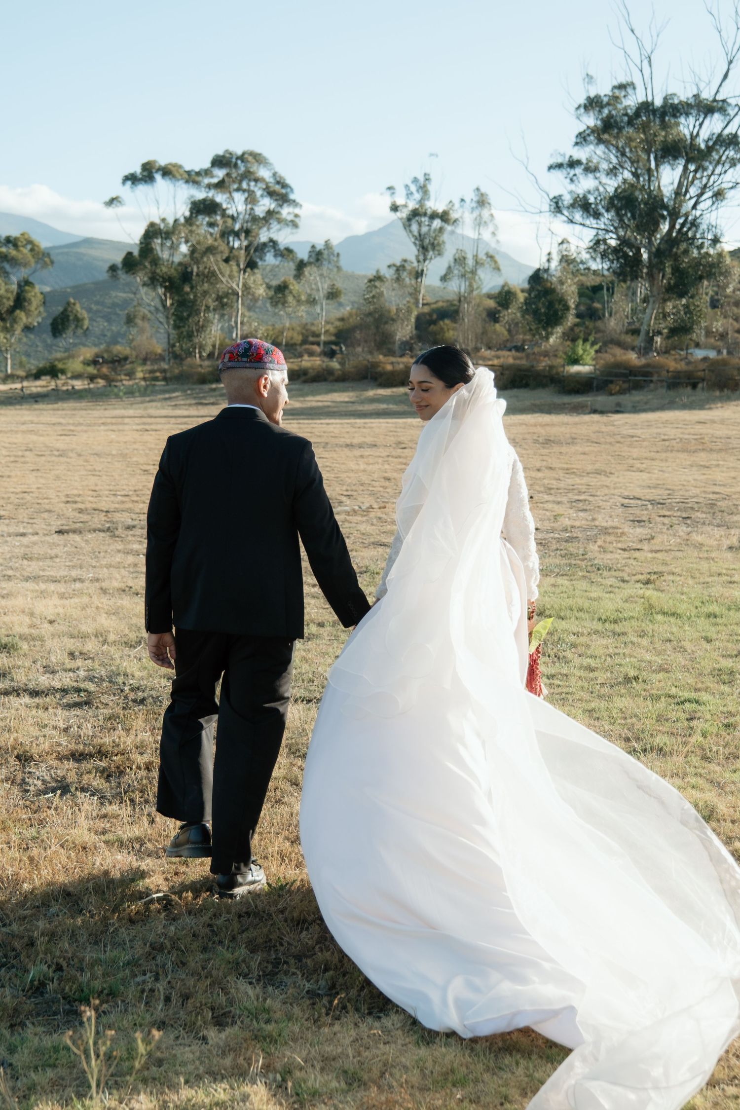 Bride in flowing white gown and veil walks hand-in-hand with groom across golden field with mountains.