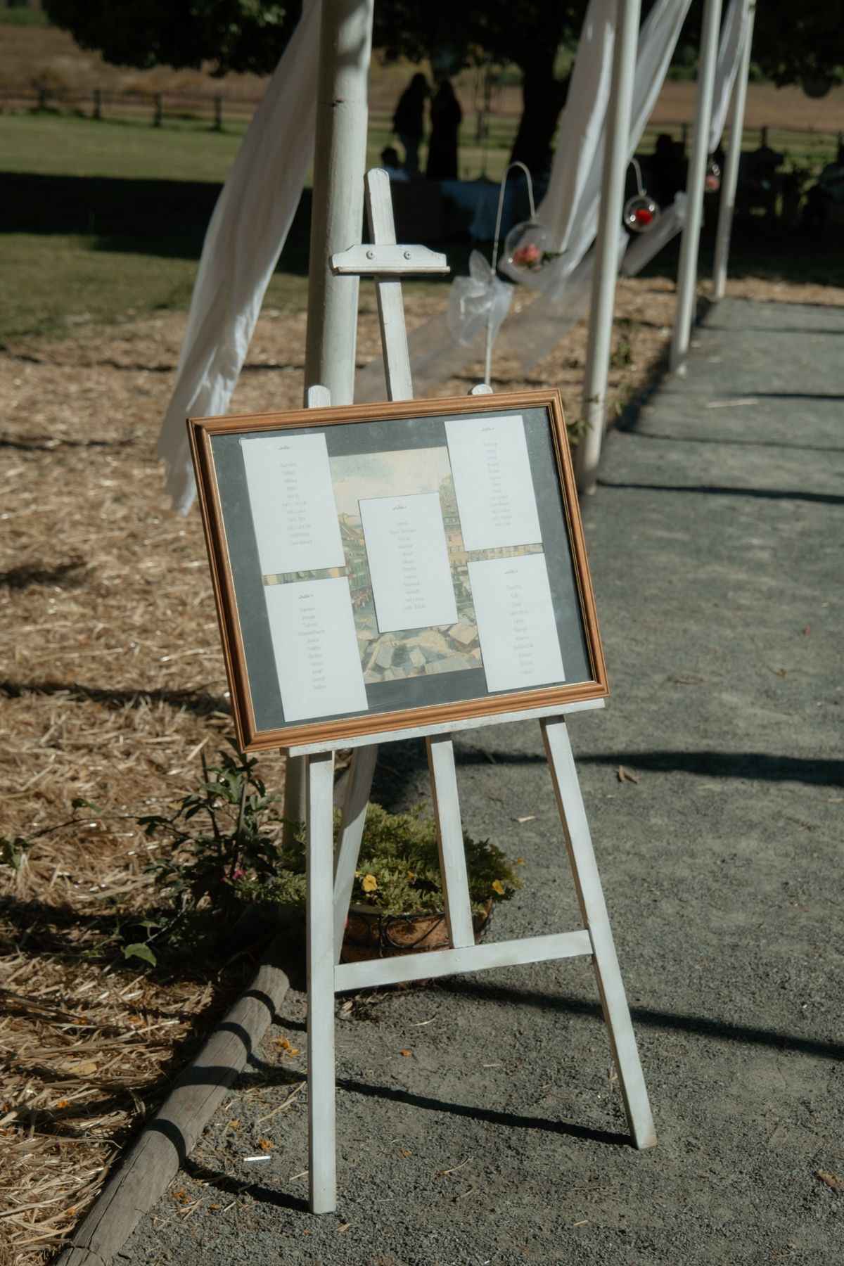 Wedding seating chart displayed in wooden frame on white easel at outdoor ceremony entrance pathway.