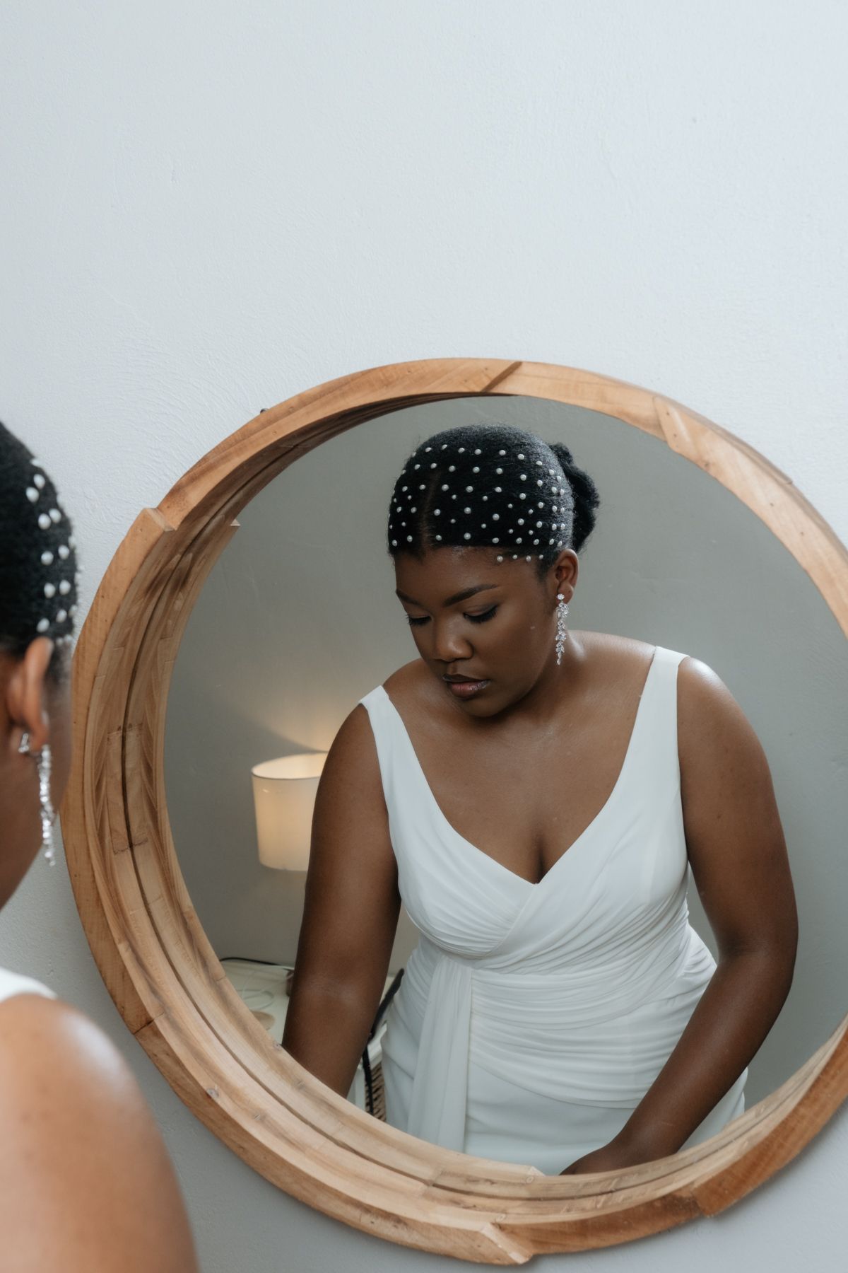 Black bride in white gown with pearl hair accessory admires her reflection in a round wooden-framed mirror.