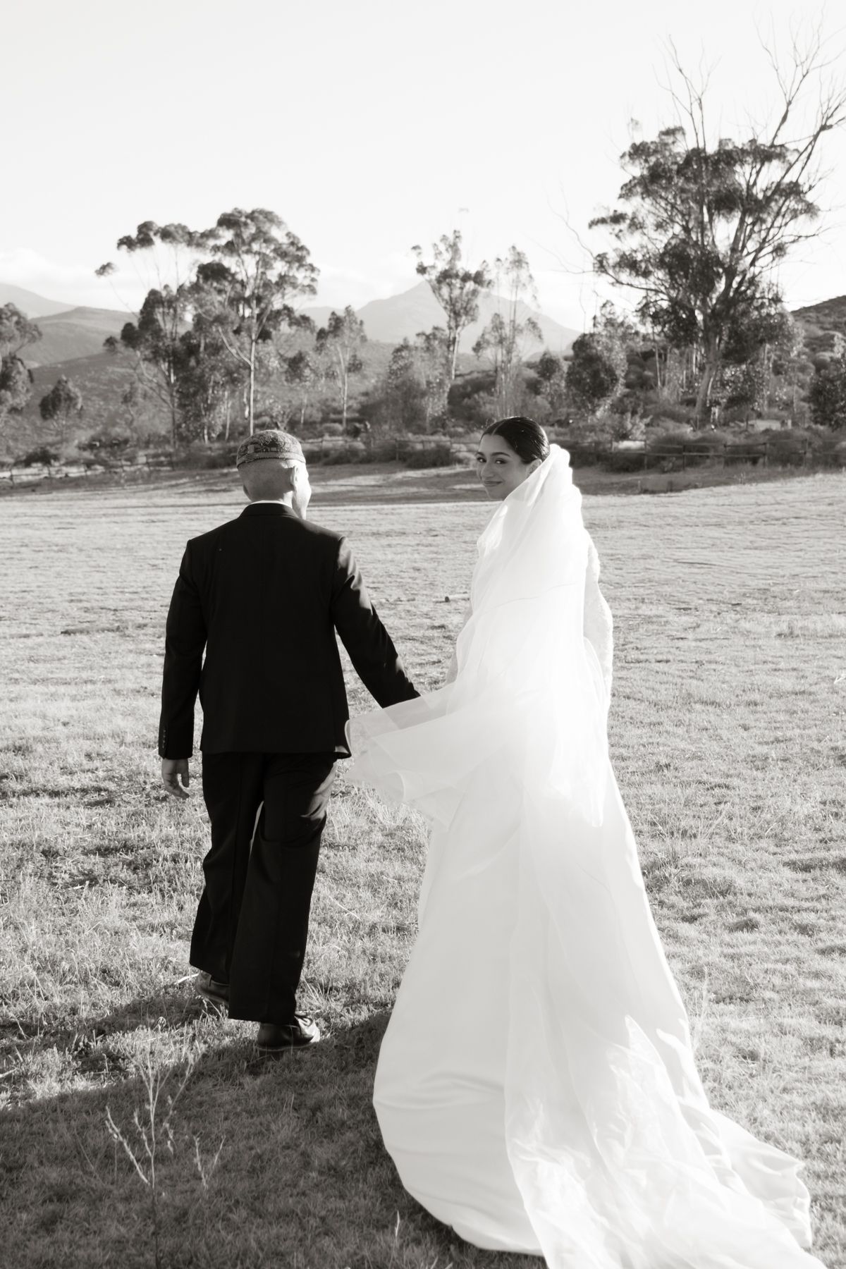 Black and white photo of bride and groom walking hand in hand across open field with mountains.