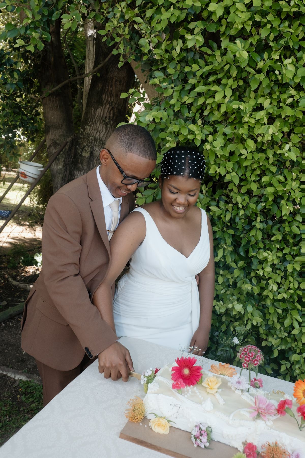 A couple cuts their wedding cake outdoors, groom in brown suit, bride in white dress with polka dot hat.