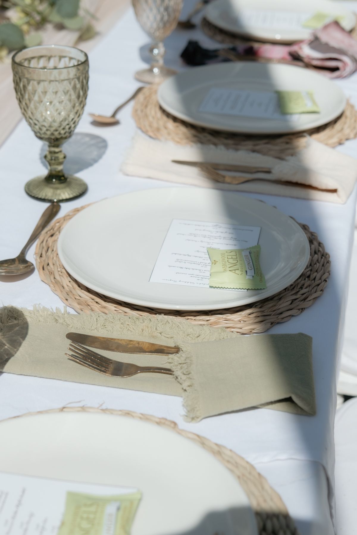 Elegant outdoor wedding table setting with woven charger plates, green goblets, and menu cards.