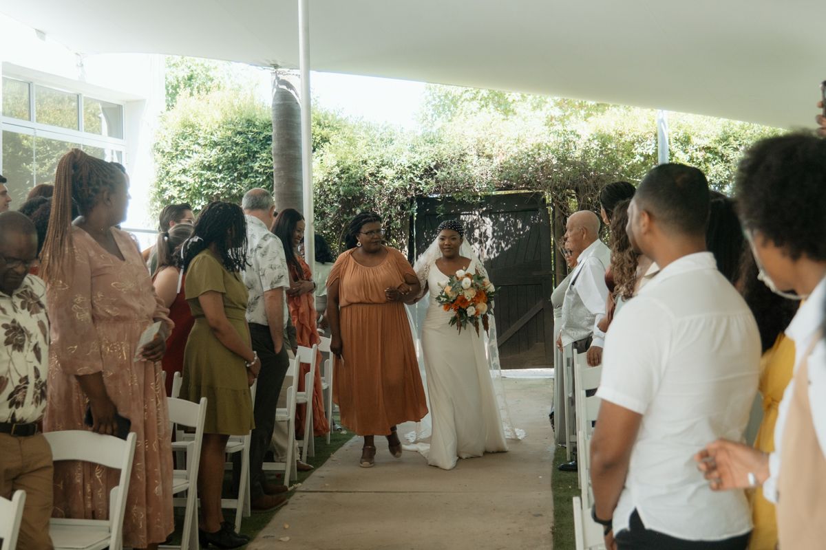 Bride walks down the aisle holding a colorful bouquet at an elegant outdoor wedding ceremony under a white tent.