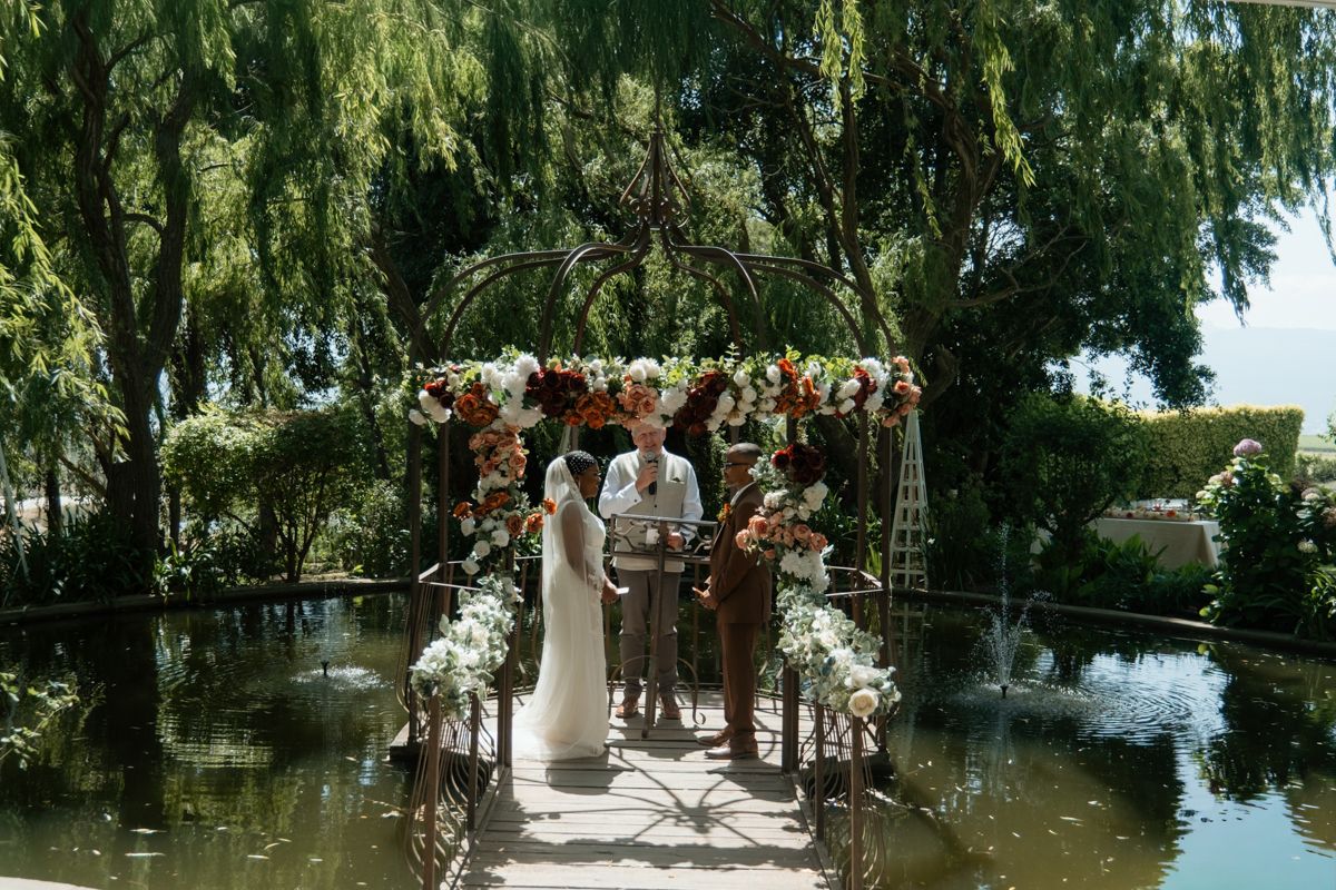 Couple exchanges vows under floral arch by tranquil pond in lush garden wedding ceremony.