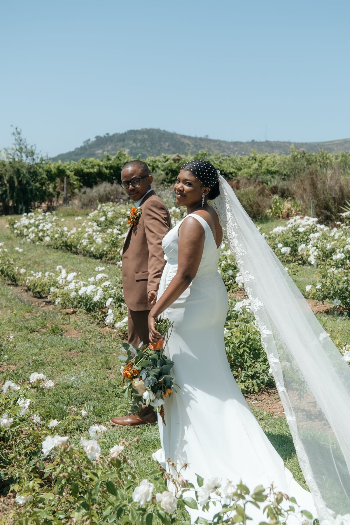 Black bride and groom pose in a lush garden with white flowers, mountains in background on their wedding day.