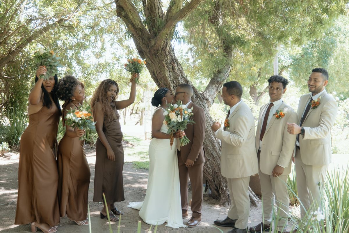 Joyful wedding party celebrating under a large tree, bridesmaids in brown dresses holding bouquets.