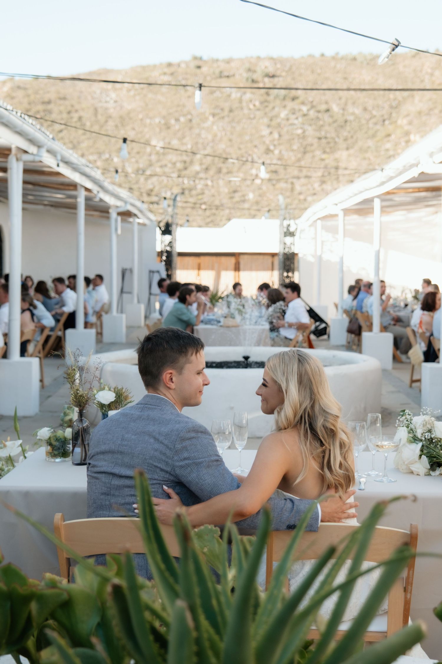 Wedding guests enjoy an outdoor reception under a white canopy with coastal views and tropical plants in the foreground.