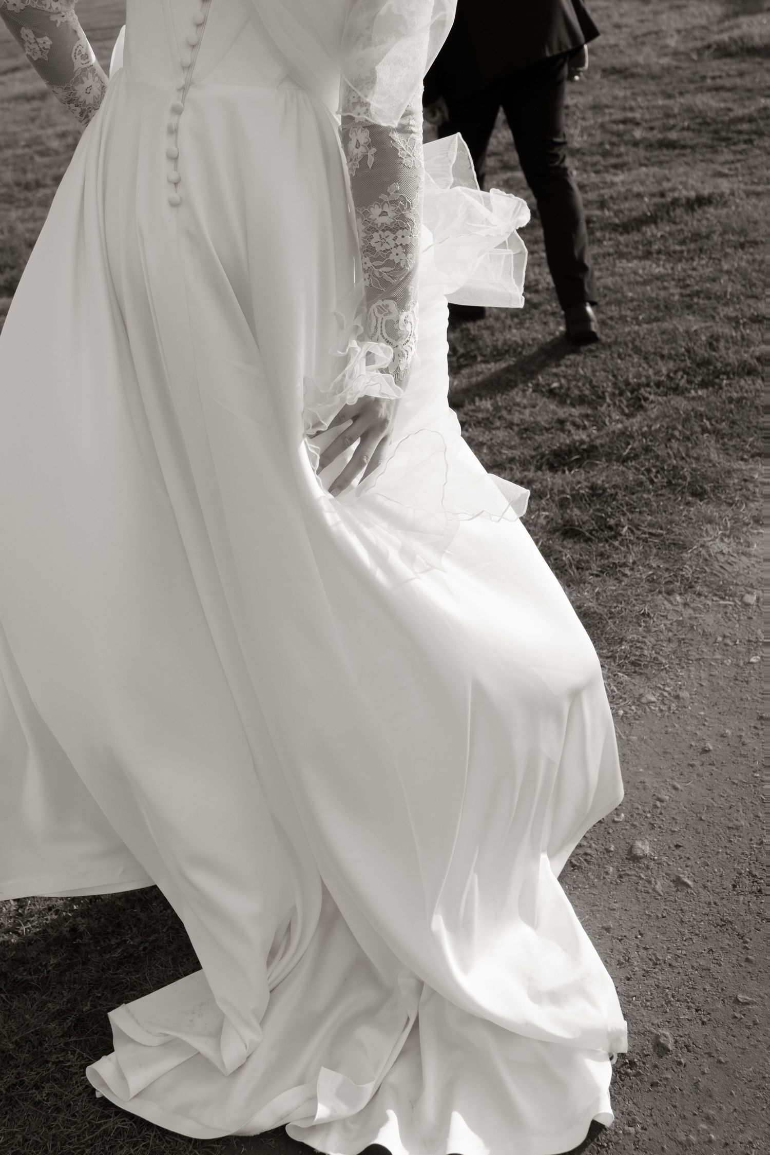 Black and white photo of a bride in a flowing lace wedding gown walking with groom on a path.