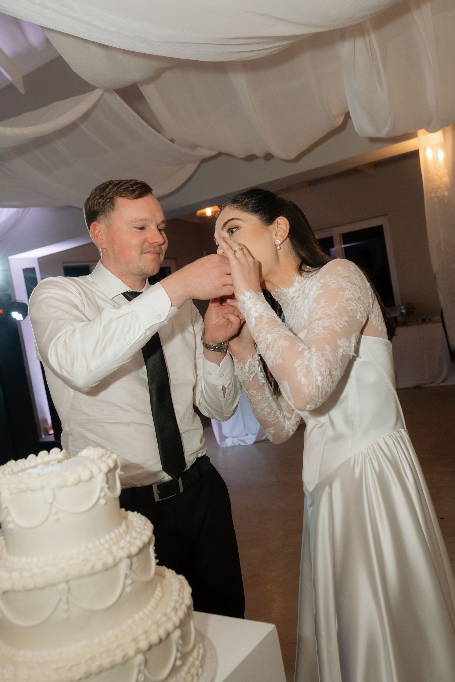 Bride and groom smiling while feeding each other wedding cake at their elegant white-decorated reception venue.