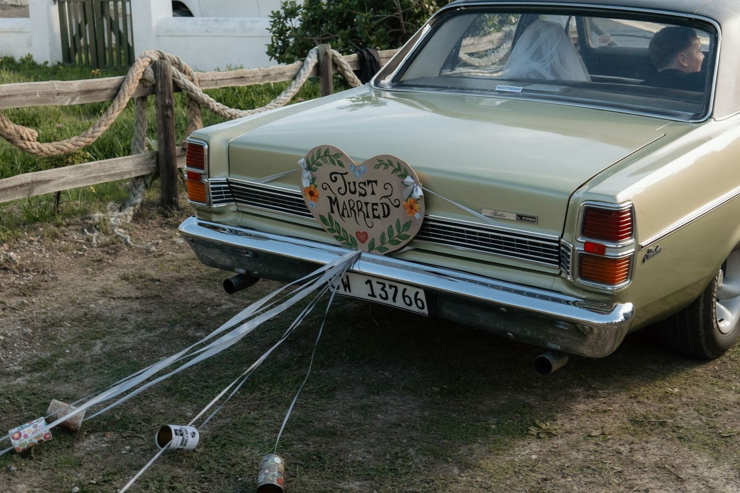 A vintage yellow car with a Just Married sign and tin cans tied to the bumper parked outdoors.
