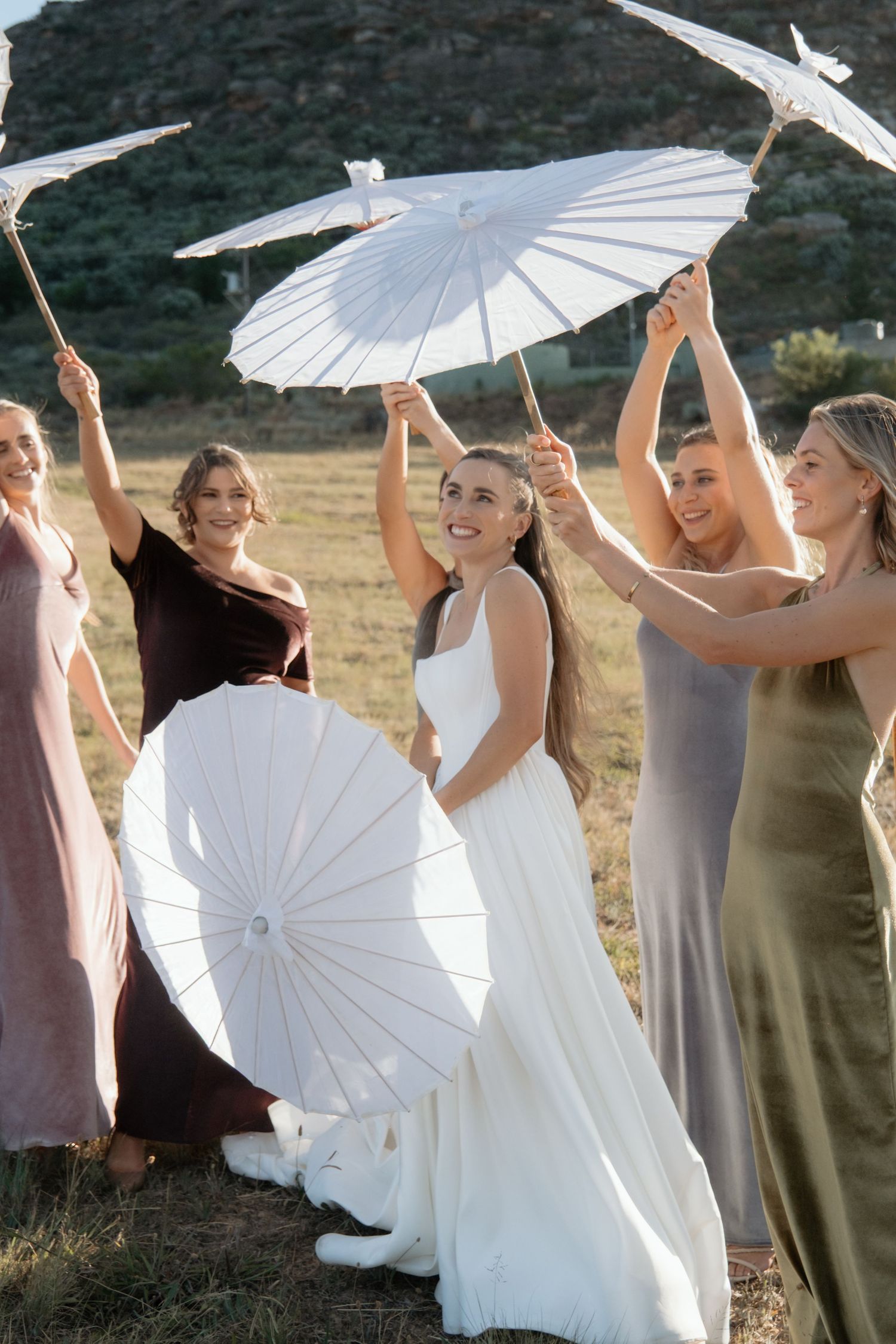 Wedding party poses outdoors with white paper parasols in a playful moment against a natural landscape.