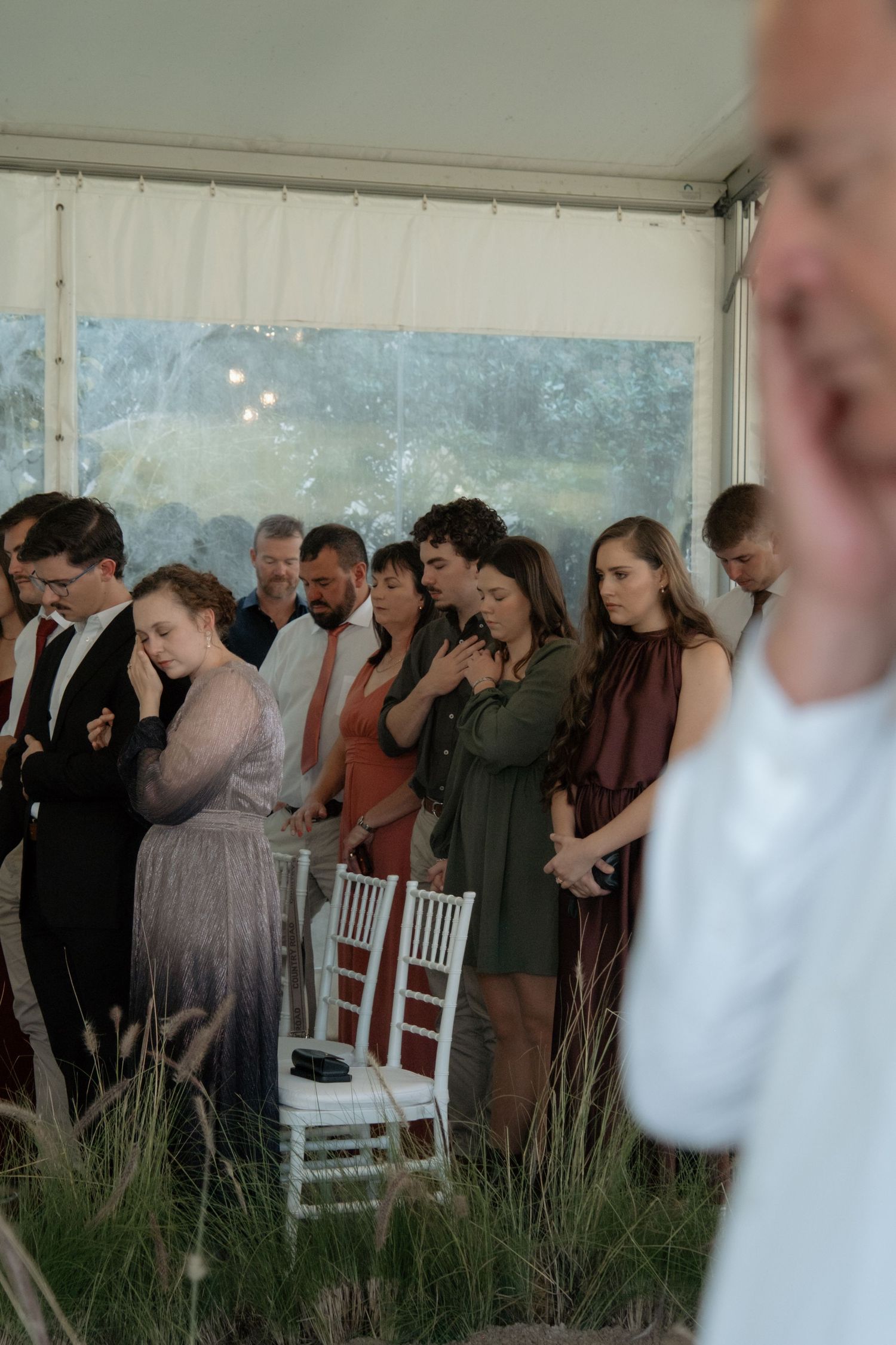 Guests standing in prayer during an outdoor wedding ceremony under a white tent with glass walls.