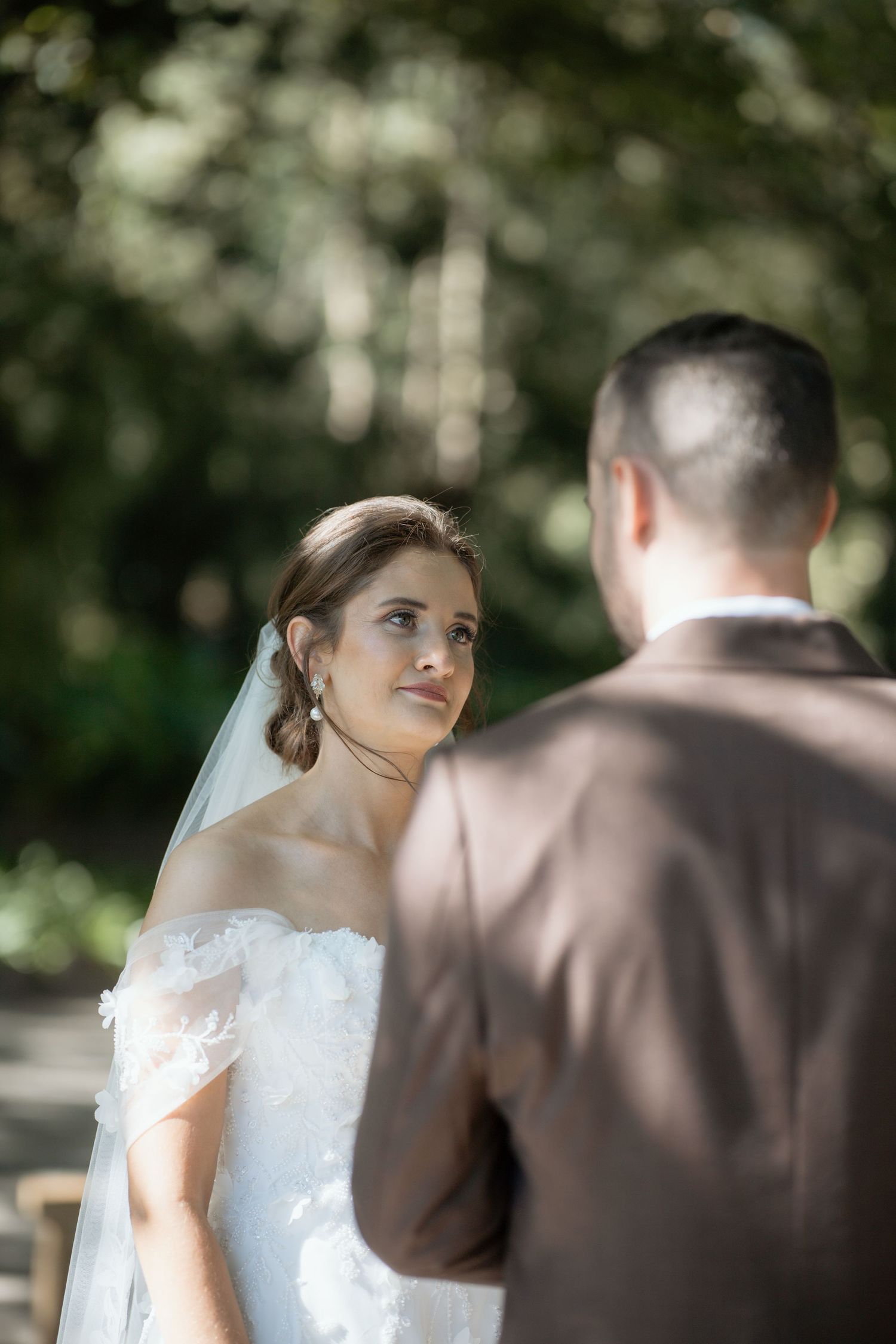 Bride in off-shoulder lace gown with veil gazes at groom during outdoor wedding ceremony.