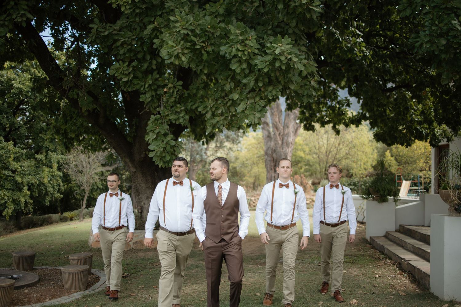 Groom and groomsmen in brown suits with suspenders walking outdoors under a large tree at a rustic wedding.