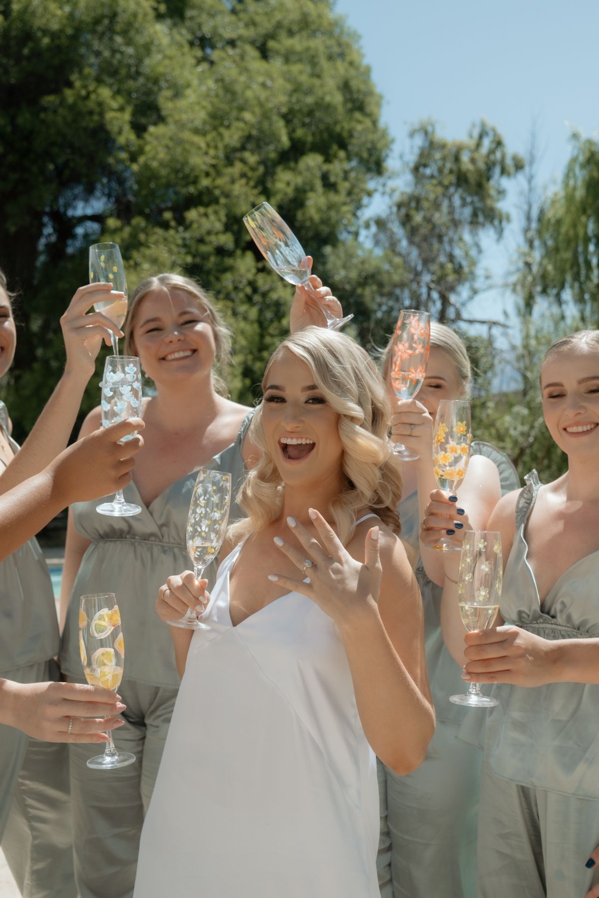 Bride showing off engagement ring while celebrating with bridesmaids holding champagne glasses outdoors.