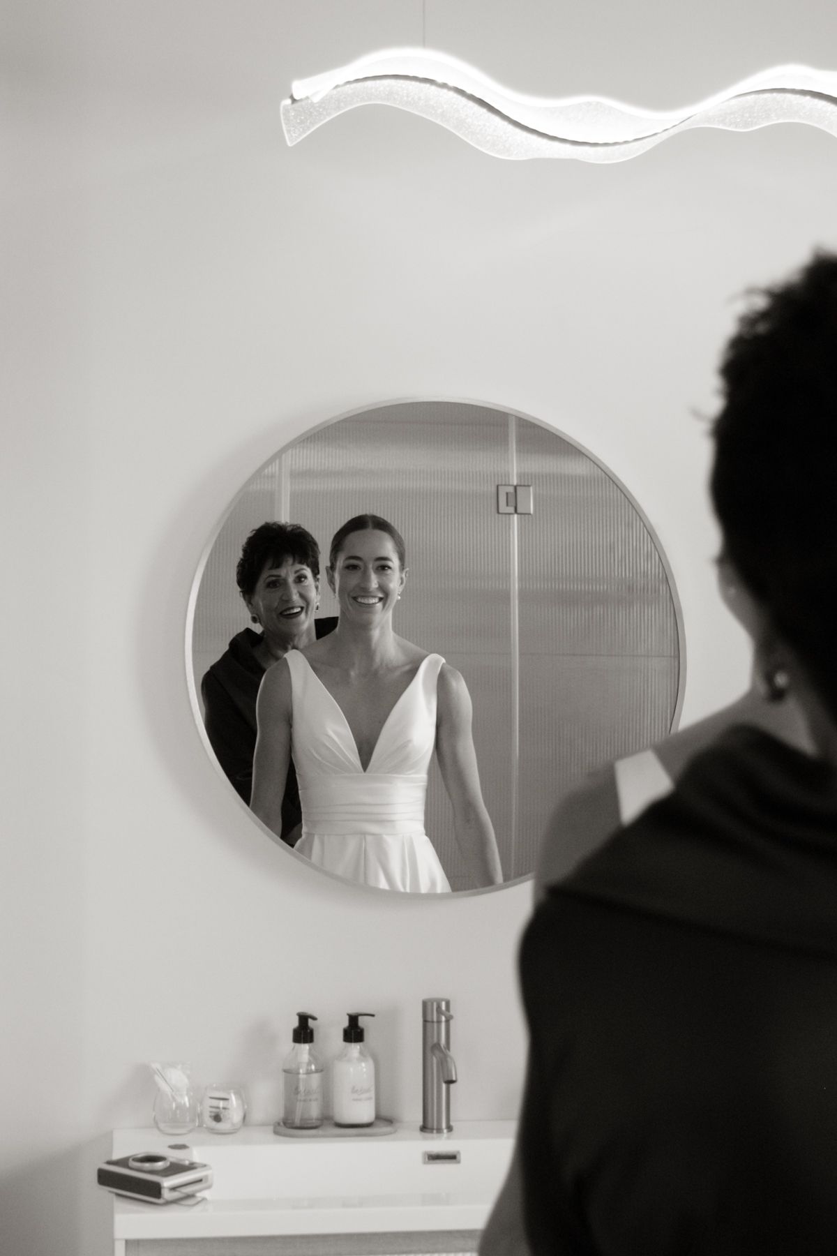 Black and white mirror reflection of a smiling bride in a white gown with a woman beside her.