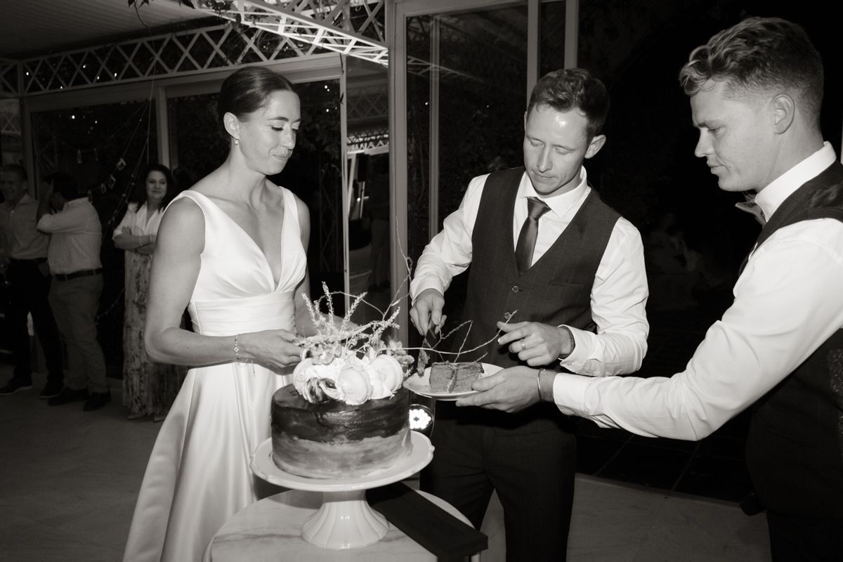 Black and white photo of bride and groom cutting a dark wedding cake assisted by a groomsman at reception.