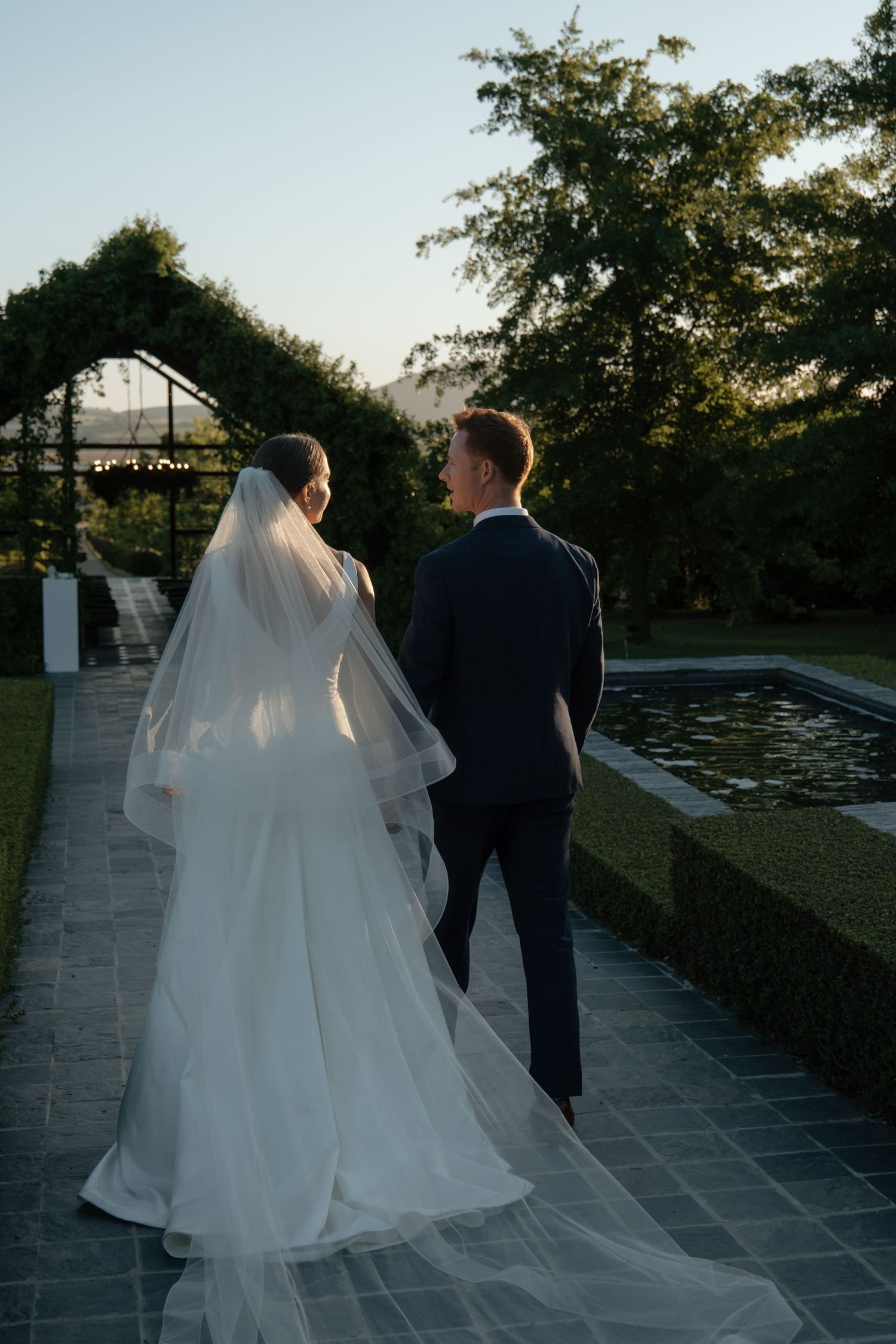 Bride and groom walking hand in hand through a lush garden venue at golden hour sunset.