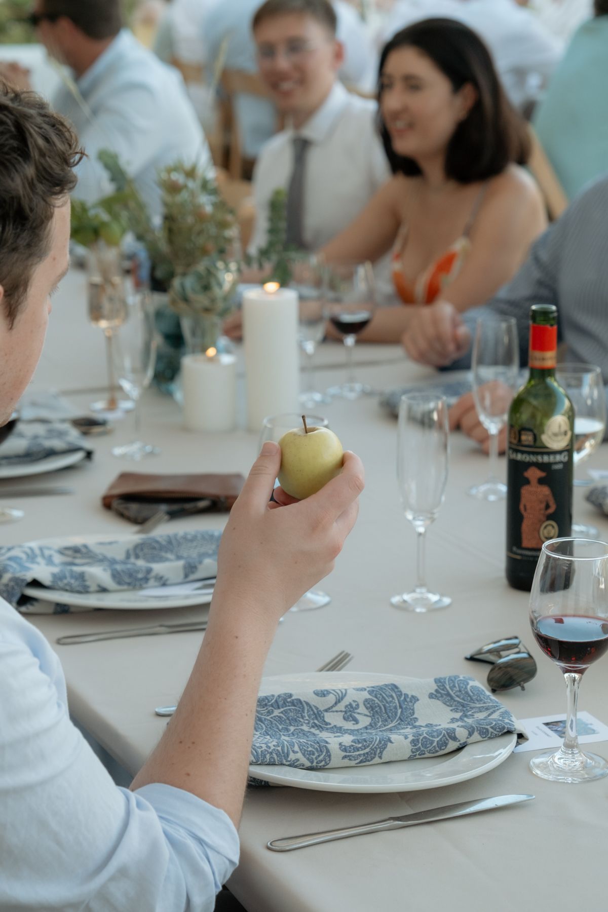 Wedding guest holding a green apple at an elegantly set reception table with candles, wine, and blue floral napkins.