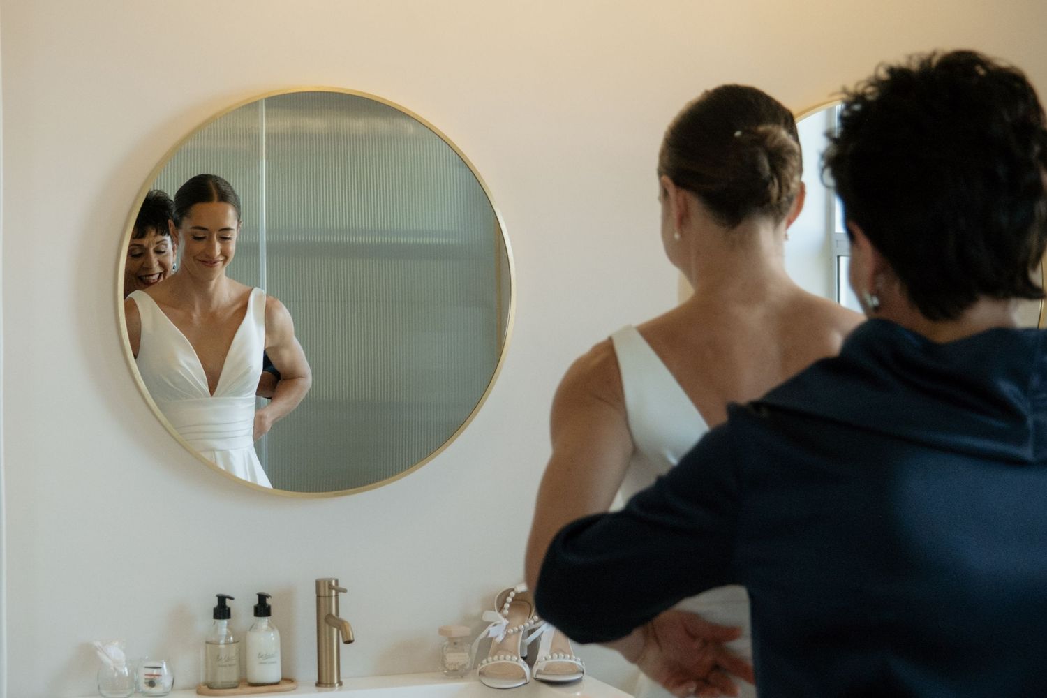 Bride in white wedding dress admiring her reflection in a round mirror while being assisted in a bathroom.