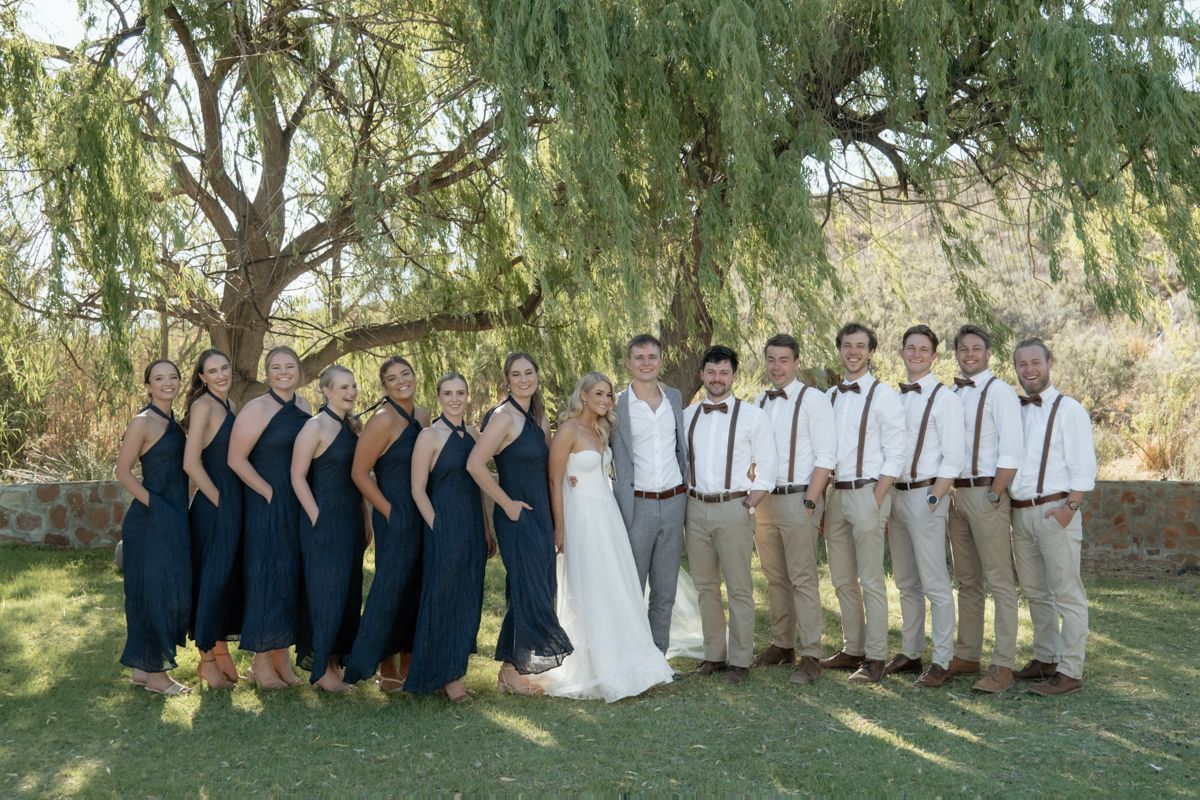 Wedding party posing under a willow tree with bridesmaids in navy dresses and groomsmen in tan pants with suspenders.