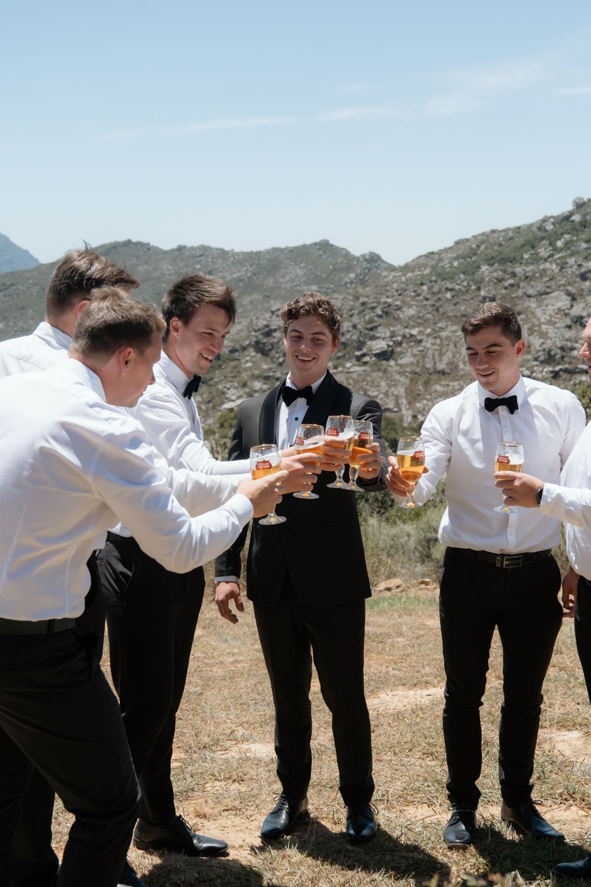 Groomsmen in black tuxedos toasting with amber drinks outdoors against a scenic mountain backdrop.