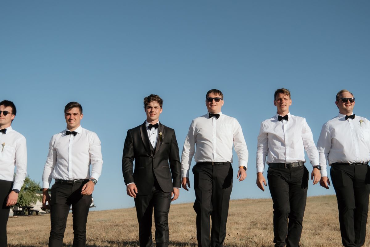 Groom and groomsmen in black tuxedos and bow ties standing in a sunny field under a clear blue sky.