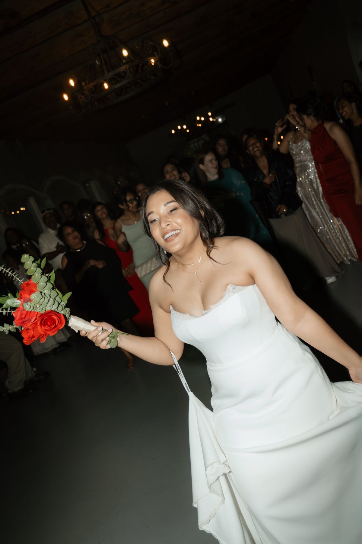 Smiling bride in strapless white gown prepares to toss red rose bouquet to guests at wedding reception.