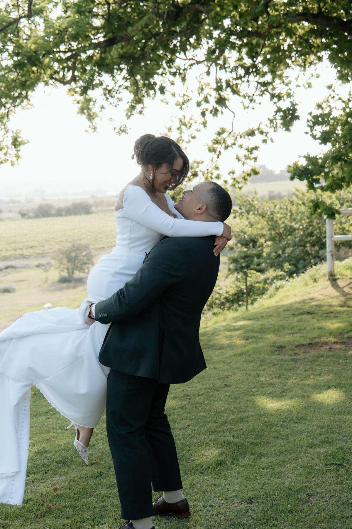 A groom lifts his smiling bride in a white wedding dress under a tree with a scenic green landscape behind them.