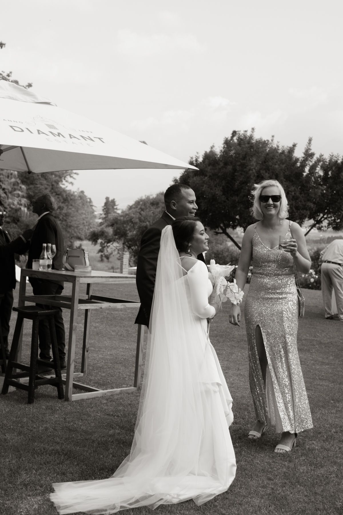 Bride in white gown and veil chats with guests at outdoor wedding reception near Diamant umbrella.