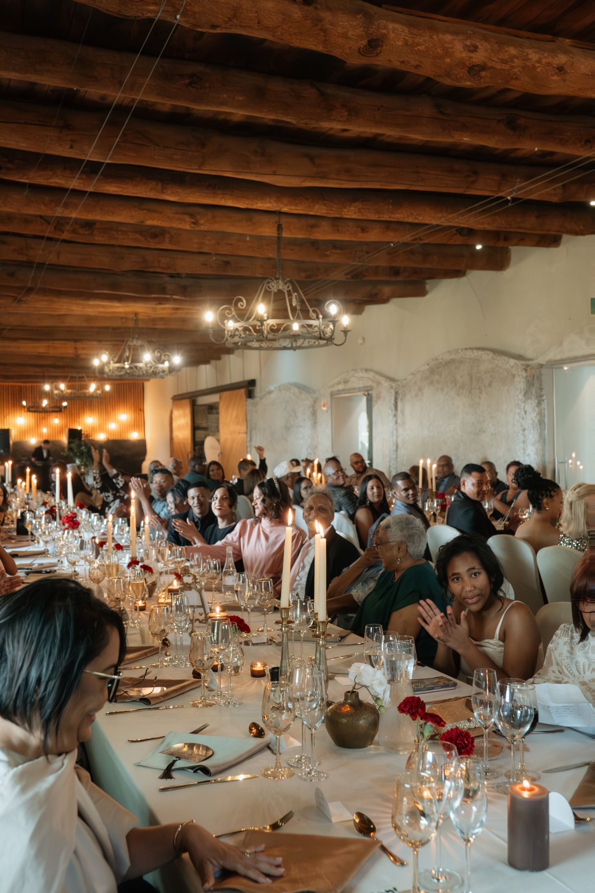 Elegant wedding reception dinner in a rustic venue with wooden ceiling, candlelit long tables and guests.