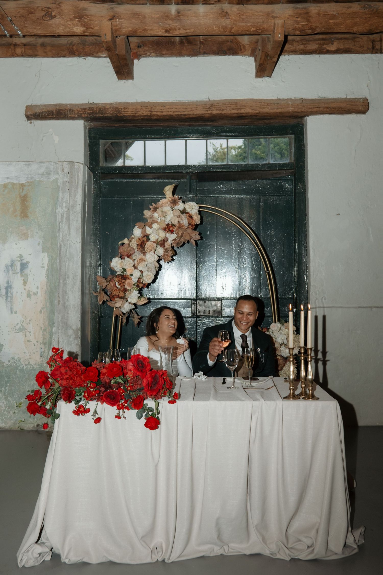 Newlyweds toast at sweetheart table adorned with red floral arrangements and gold candelabras in rustic venue.