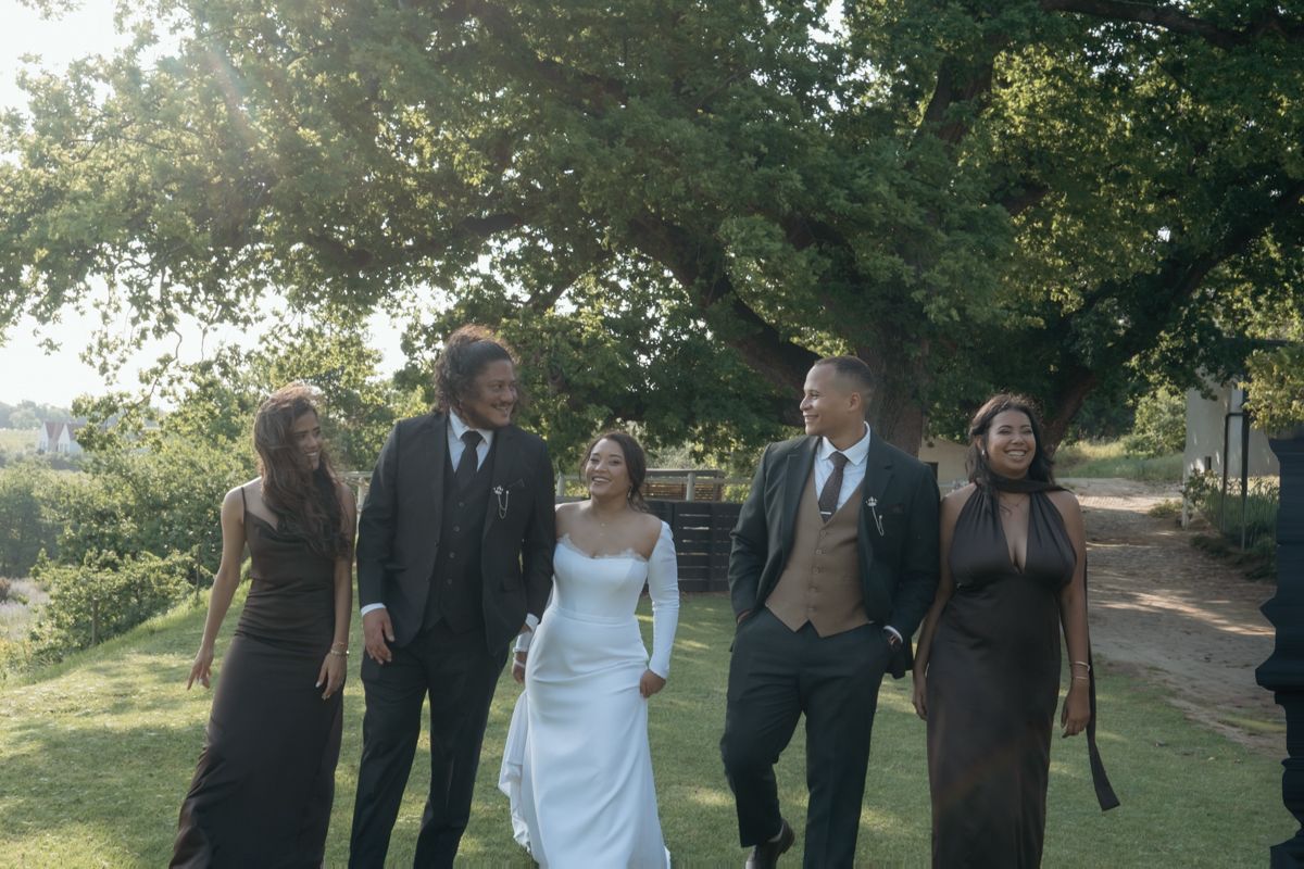Bride in white gown walks with wedding party in dark formal attire under a large tree outdoors.