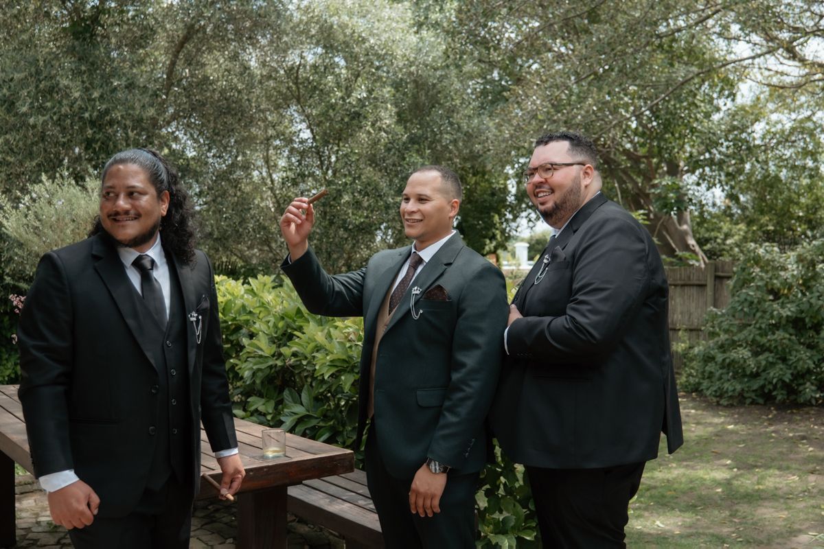 Three groomsmen in dark suits laughing and gesturing outdoors among trees before the wedding ceremony.