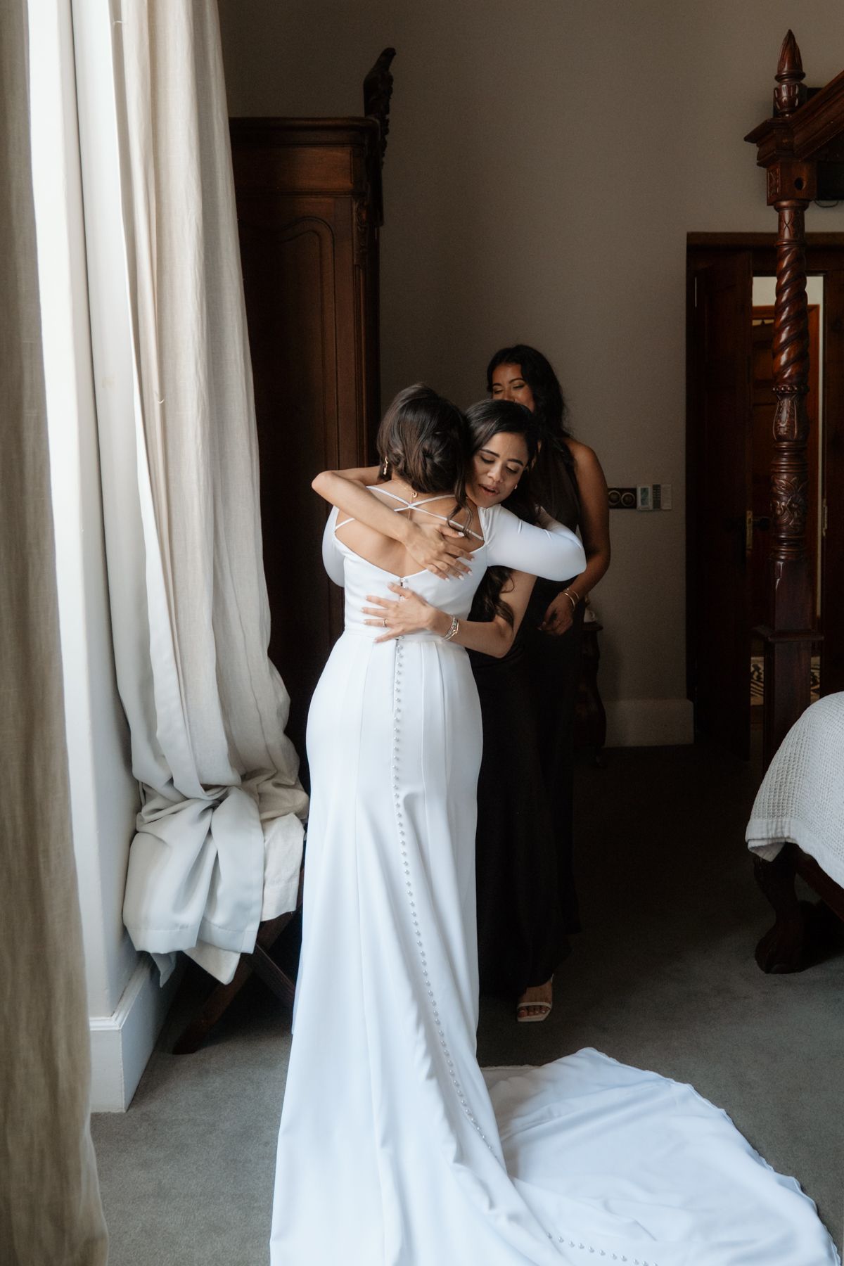 Bride in fitted white button-back wedding gown embracing bridesmaid in elegant hotel suite.