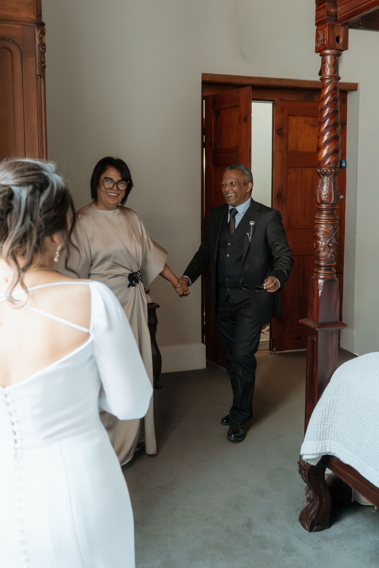 Bride in white gown sees parents entering room during wedding day preparations.