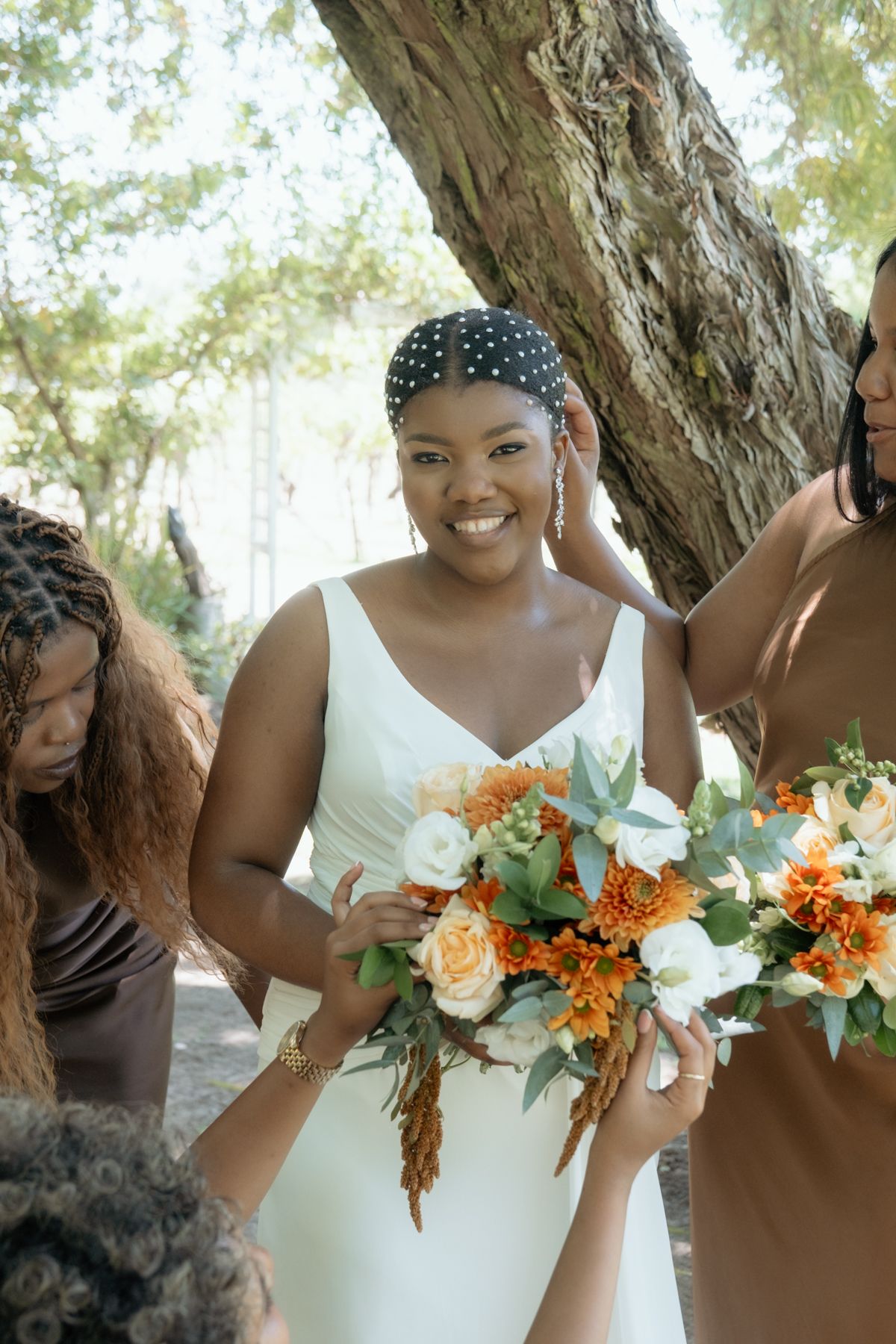 Radiant bride in white dress smiles while holding orange and white bouquet surrounded by bridesmaids in brown dresses.