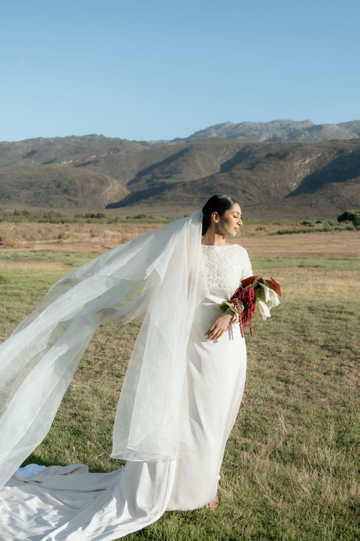 Bride in white long-sleeve gown and flowing veil holds red bouquet in open field with mountain landscape behind her.