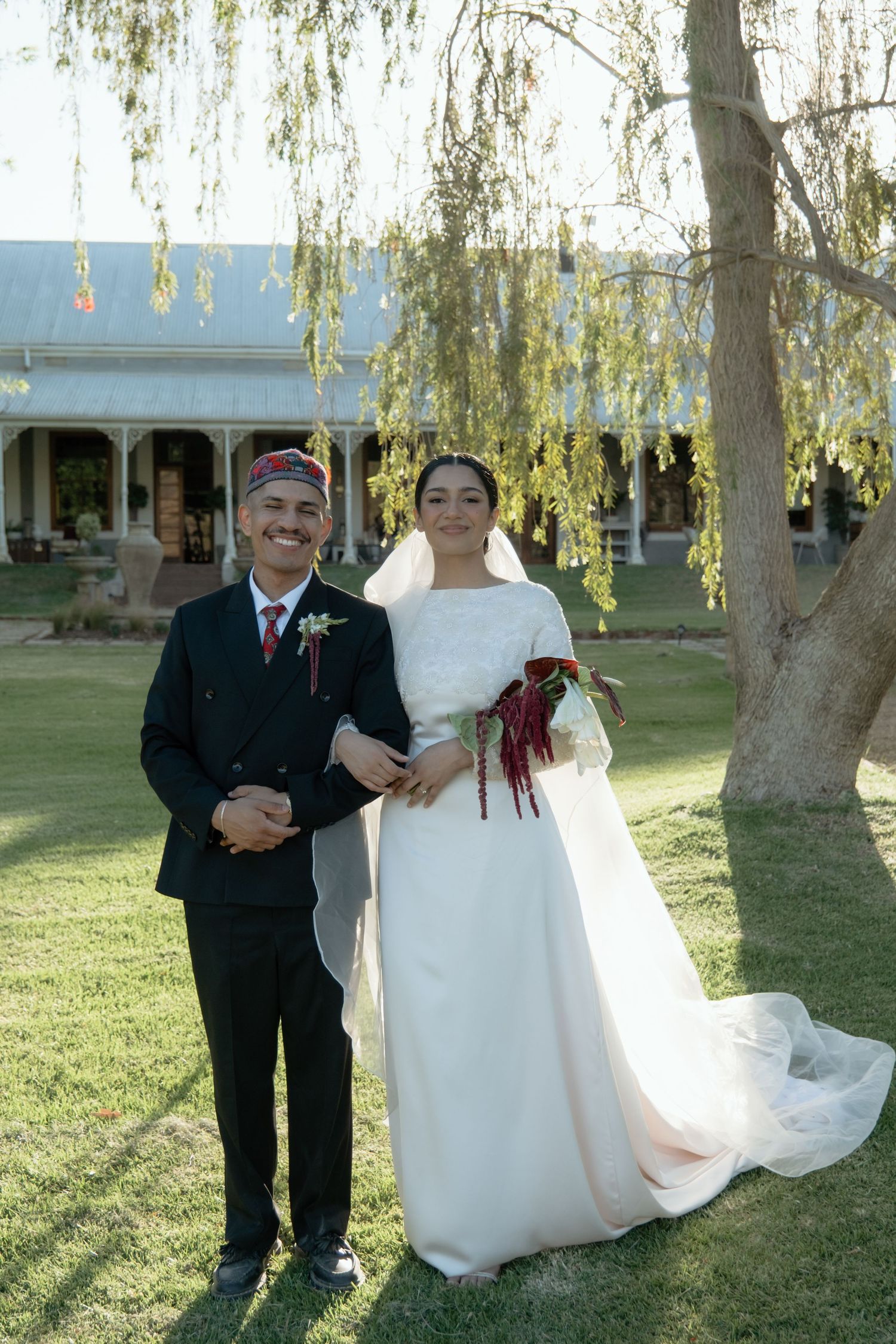 Bride in elegant white gown with bouquet poses with groom in black suit and kippah at outdoor wedding.