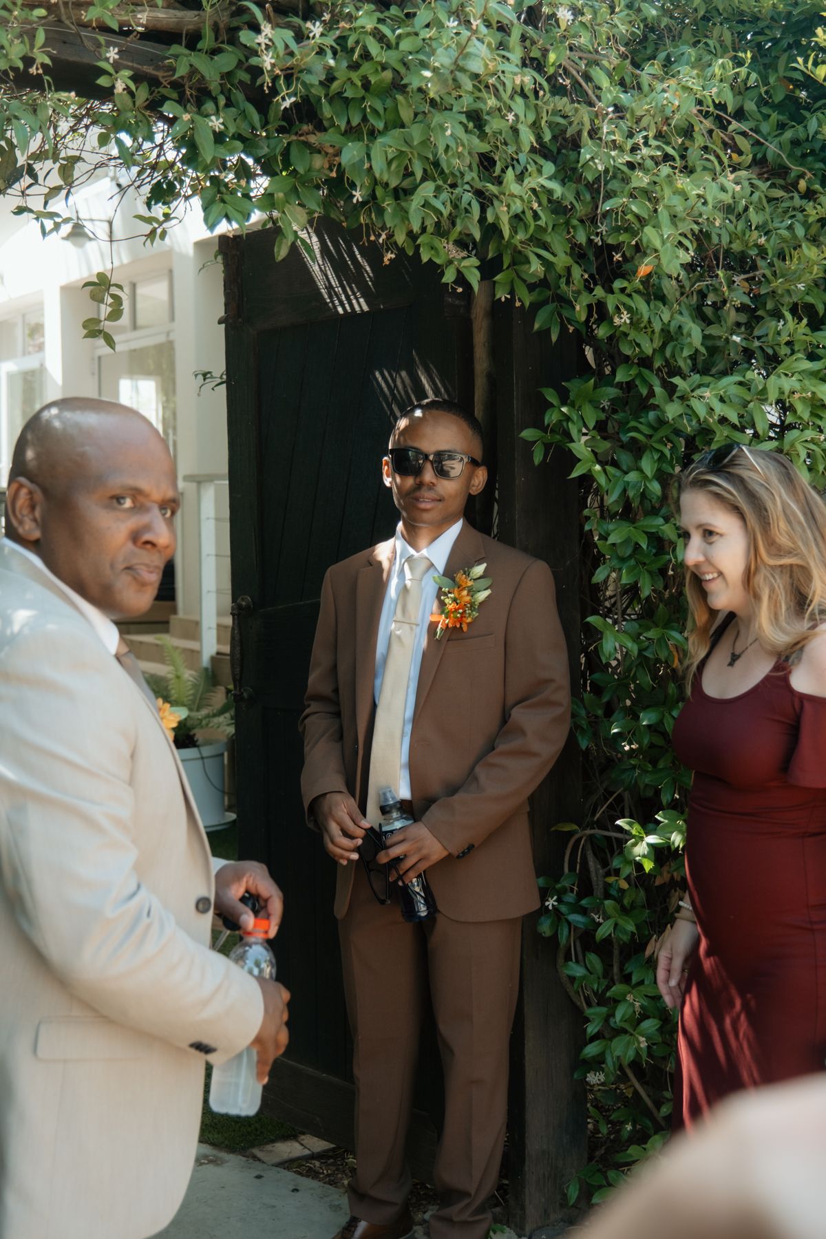 Groom in brown suit with floral boutonniere stands with guests near ivy-covered entrance at outdoor wedding.