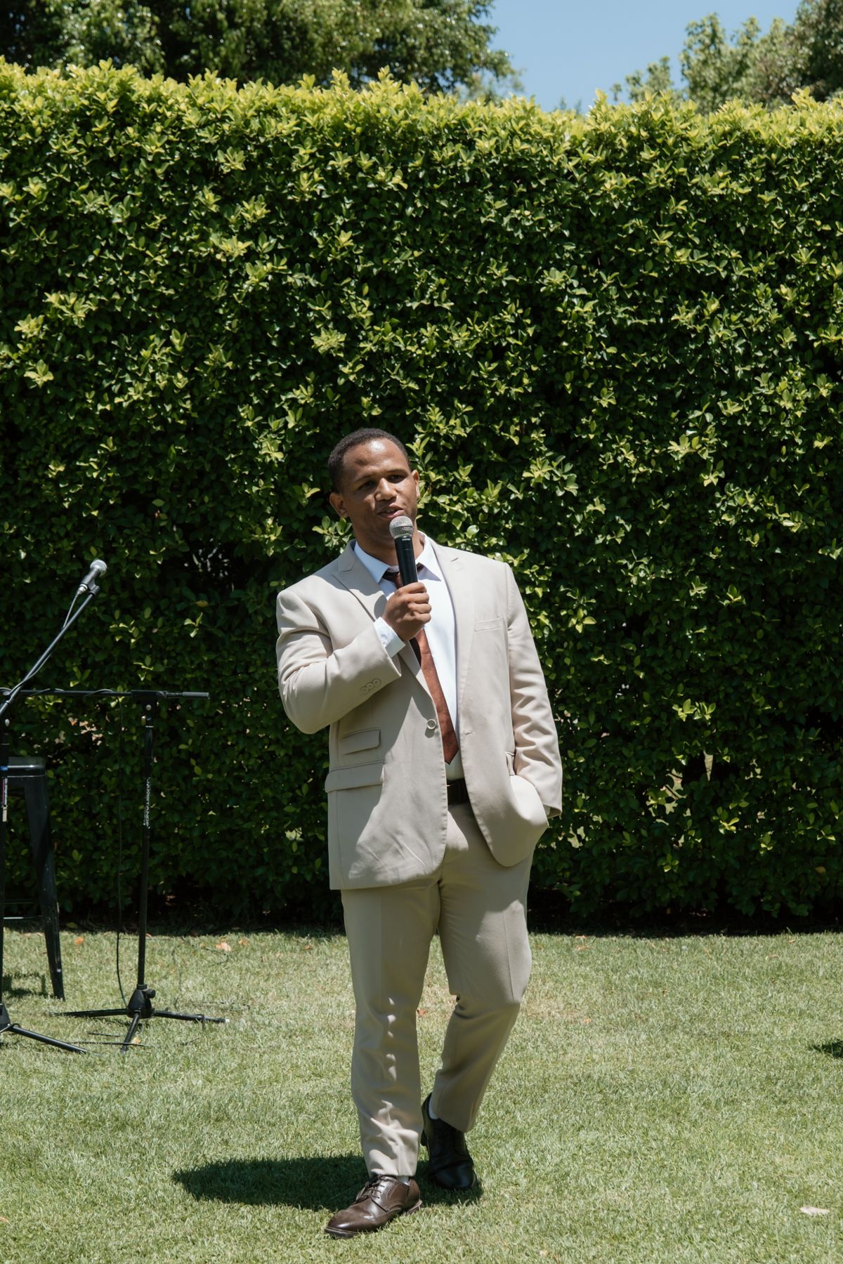 Man in beige suit speaking into microphone at outdoor wedding reception on green lawn.