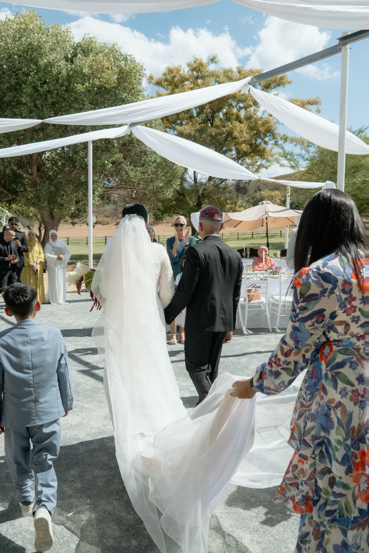 Bride walking down the aisle in a white gown and veil at an outdoor wedding ceremony.